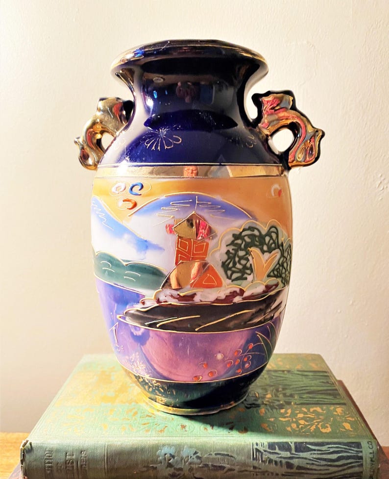 May include: A vintage, hand-painted ceramic vase with a dark blue top and gold accents. The body features a landscape scene with a red building, mountains, and trees. The vase has two ornate handles and sits on a green book.