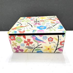 May include: A rectangular storage box with a clear lid featuring a colorful pattern of birds, flowers, and leaves in shades of red, blue, yellow, pink, and green. The box has a small, clear, square knob on the lid and a black interior lining.