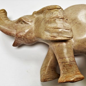 May include: A hand-carved stone elephant figurine with a light brown color and a textured surface. The elephant has its trunk raised and its head tilted to the side.