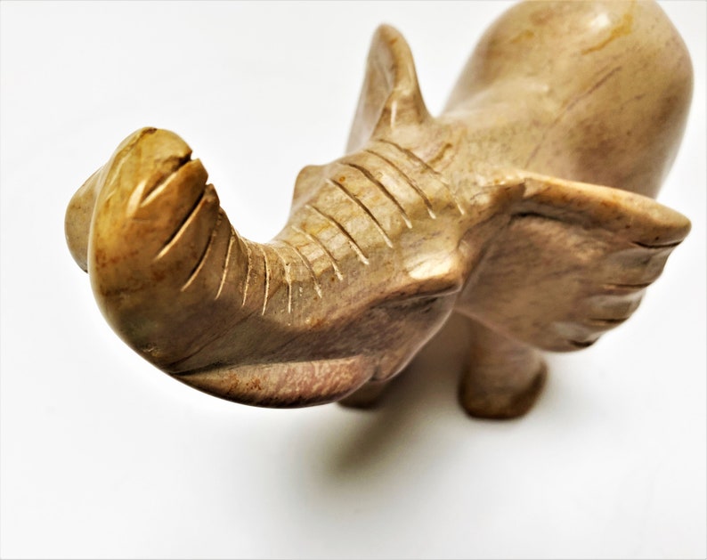 May include: A hand-carved stone elephant figurine with a light brown color. The elephant's trunk is raised and curled, and its ears are spread out.
