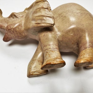 May include: A hand-carved stone elephant figurine with a light brown and pink color. The elephant is standing with its trunk raised and its head tilted to the side.