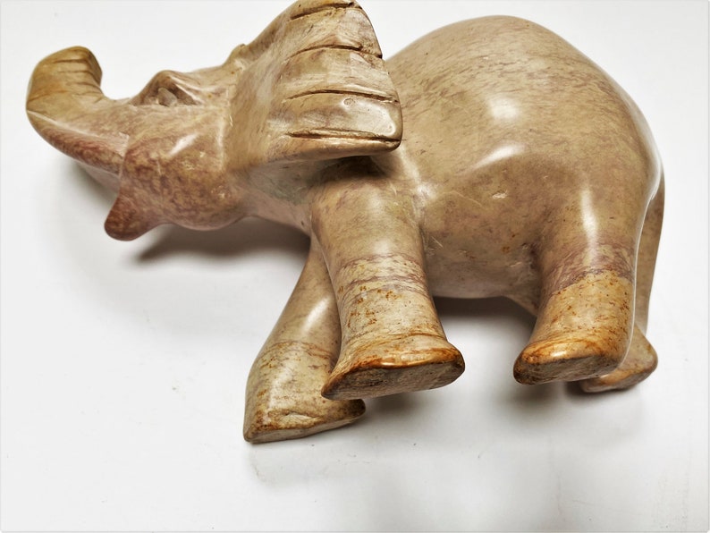 May include: A hand-carved stone elephant figurine with a light brown and pink color. The elephant is standing with its trunk raised and its head tilted to the side.