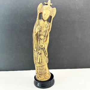 May include: A gold-colored figurine of a robed figure with ornate details, standing on a black circular base. The figure has a detailed headdress and holds objects in both hands. The background is a gradient of black and white.