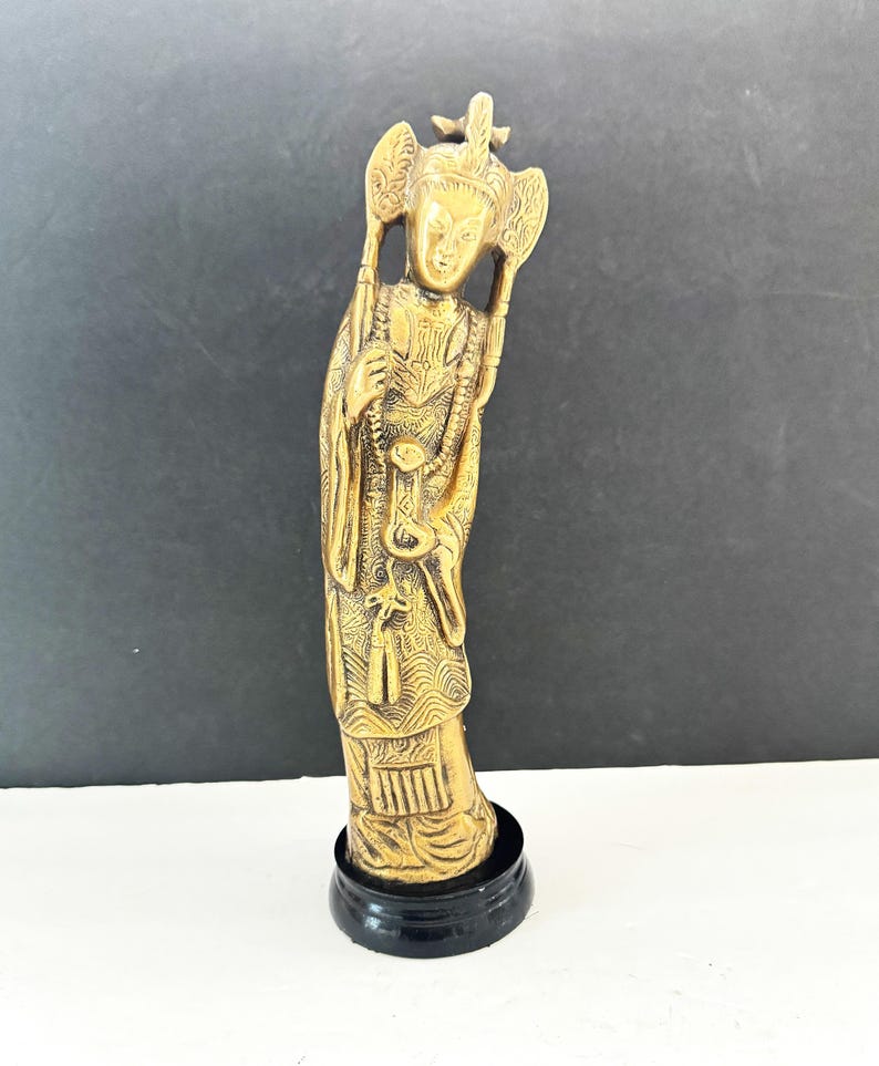 May include: A gold-colored figurine of a robed figure with ornate details, standing on a black circular base. The figure has a detailed headdress and holds objects in both hands. The background is a gradient of black and white.