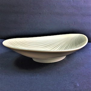May include: An oval-shaped ceramic bowl in a pale cream color. The interior surface features a series of raised, radiating lines that extend from the center toward the rim, creating a textured, fan-like pattern across the entire basin.