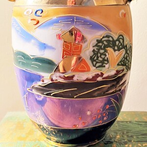 May include: A decorative ceramic vase with a landscape design. The vase features a mountain scene with a building, trees, and water, painted in shades of blue, purple, orange, and gold. The vase has a gold rim and base.