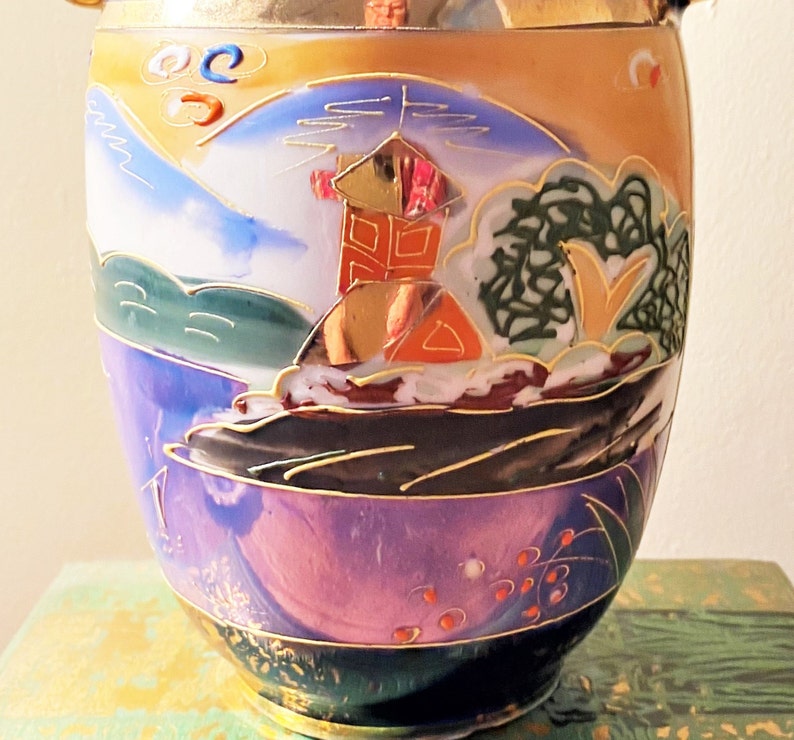 May include: A decorative ceramic vase with a landscape design. The vase features a mountain scene with a building, trees, and water, painted in shades of blue, purple, orange, and gold. The vase has a gold rim and base.
