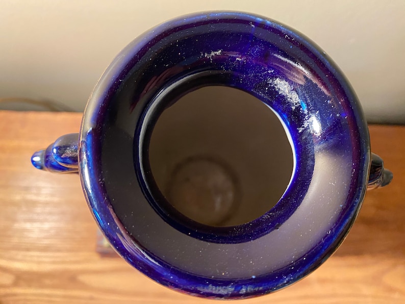 May include: A cobalt blue ceramic vase with a wide opening and two decorative handles. The vase has a glossy finish and a light interior. The top-down perspective highlights the vase's shape and color.