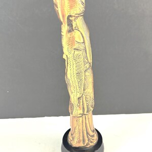 May include: A detailed, golden-colored statue of a figure with ornate clothing and a headdress, mounted on a black circular base. The statue has intricate carvings and a smooth finish, showcasing a classical design.