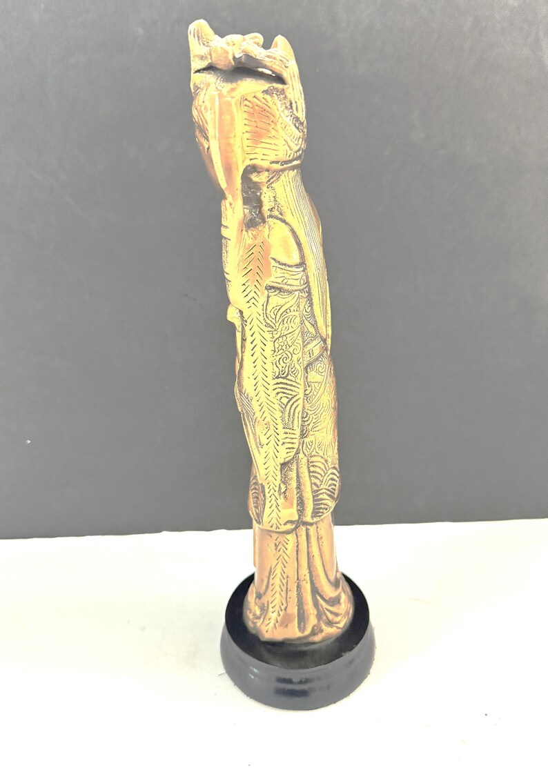May include: A detailed, golden-colored statue of a figure with ornate clothing and a headdress, mounted on a black circular base. The statue has intricate carvings and a smooth finish, showcasing a classical design.