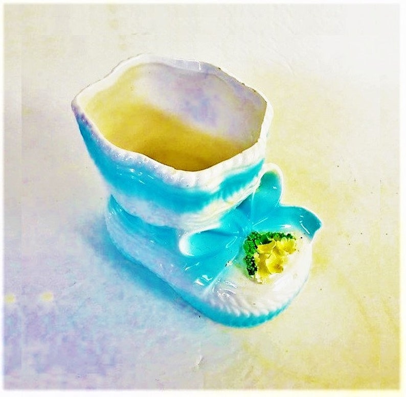 May include: A ceramic shoe-shaped planter featuring a glossy white and bright blue finish with an iridescent sheen. The front of the shoe is decorated with a small cluster of moulded yellow flowers and green leaves, with a blue bow detail.