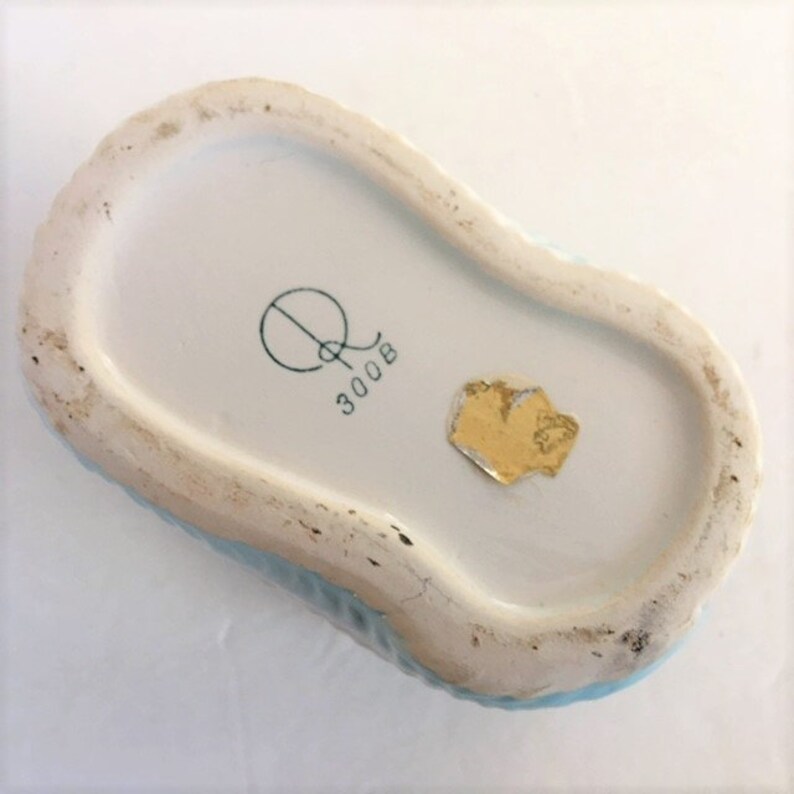May include: The underside of a ceramic object featuring a light blue textured exterior. The base is white with a printed mark consisting of a stylised letter Q above the number 300B. A small, partially torn gold-coloured foil sticker is also present.