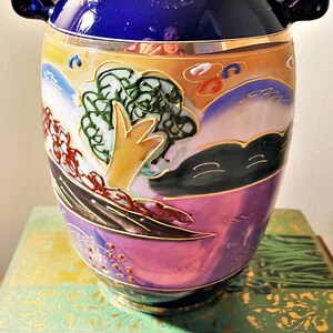 May include: A decorative ceramic vase with a vibrant design. The vase features a deep blue top, a gold rim, and a body adorned with a landscape scene in shades of purple, yellow, green, and brown. The vase has two small handles.