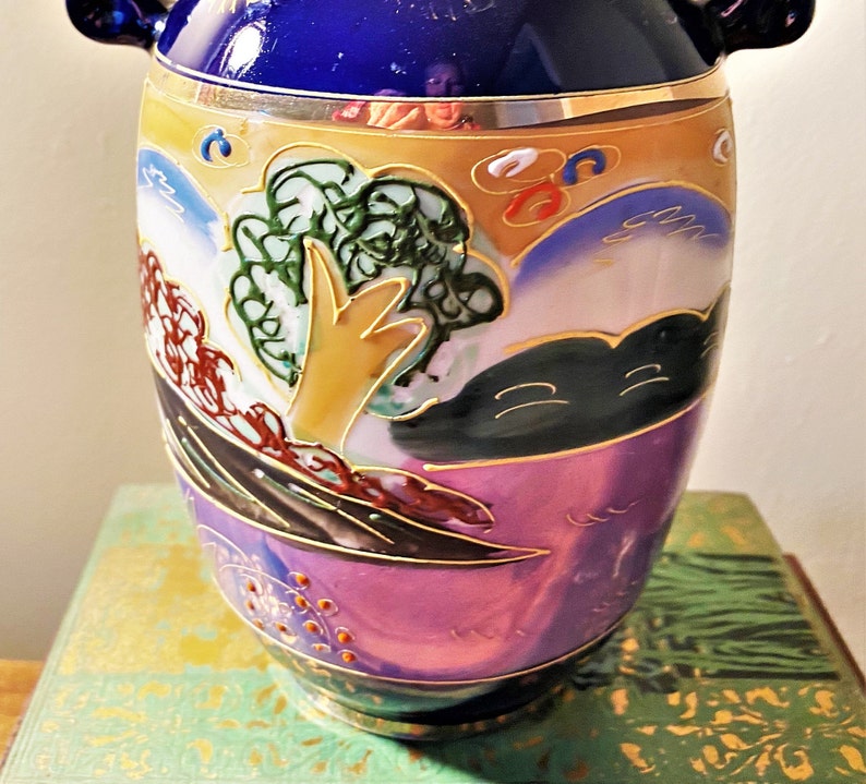 May include: A decorative ceramic vase with a vibrant design. The vase features a deep blue top, a gold rim, and a body adorned with a landscape scene in shades of purple, yellow, green, and brown. The vase has two small handles.