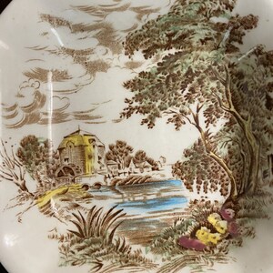 Pode incluir: Um prato de cer&acirc;mica vintage com uma cena de paisagem marrom e verde. A cena representa uma pequena cabana com uma ponte sobre um rio azul, cercada por &aacute;rvores e nuvens.