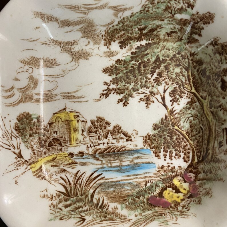 Pode incluir: Um prato de cer&acirc;mica vintage com uma cena de paisagem marrom e verde. A cena representa uma pequena cabana com uma ponte sobre um rio azul, cercada por &aacute;rvores e nuvens.