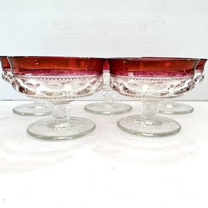 May include: Set of six vintage clear glass dessert bowls with ruby red rims. The bowls have a decorative pattern around the sides and sit on a clear pedestal base. The bowls are ideal for serving desserts.