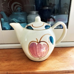 May include: A white ceramic teapot with a pink apple design and blue leaves. The teapot has a lid and a handle.