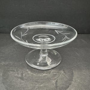 May include: A clear glass pedestal dish with a round base and a shallow, oval-shaped bowl. The bowl has etched leaf designs. The dish is elevated by a short stem. The glass is transparent, allowing light to pass through.