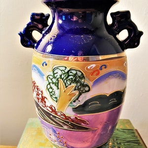 May include: A decorative ceramic vase with a dark blue top and handles. The vase features a landscape design with trees, mountains, and clouds in shades of gold, green, and purple. Gold trim accents the top and base.