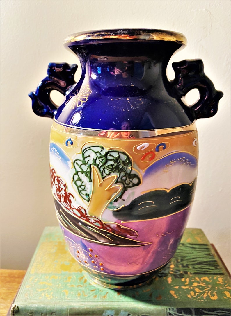 May include: A decorative ceramic vase with a dark blue top and handles. The vase features a landscape design with trees, mountains, and clouds in shades of gold, green, and purple. Gold trim accents the top and base.