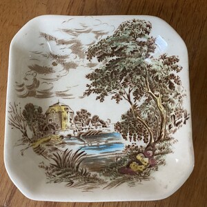Pode incluir: Um prato de servir de cer&acirc;mica branca com um design de paisagem marrom e verde. O prato apresenta uma pequena cabana, um rio, &aacute;rvores e nuvens.