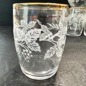 May include: Clear drinking glasses with a gold rim and white grape vine design. The glasses are cylindrical and appear to be vintage. Several glasses are visible, with one in focus. The glasses are on a dark surface.
