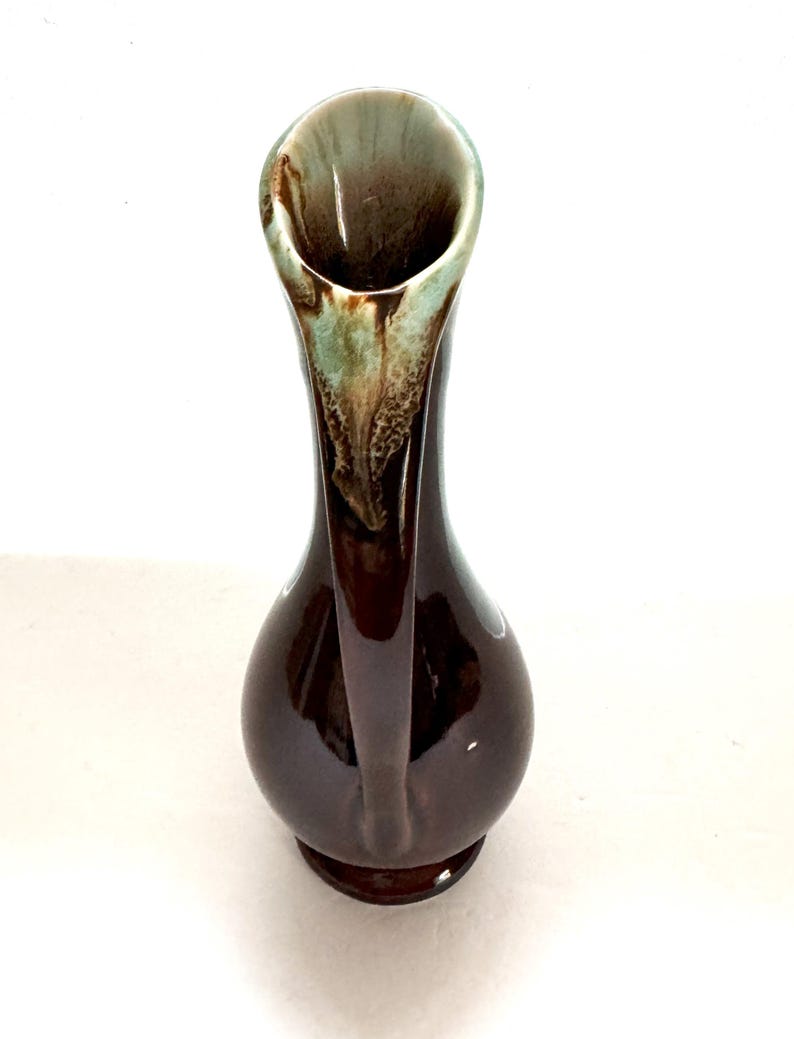 May include: A brown ceramic vase with a flared opening and a green and brown glaze. The vase has a unique shape with a curved neck and a rounded base.