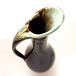 May include: A brown and green ceramic pitcher with a wide, flared opening and a curved handle. The pitcher has a mottled glaze with shades of brown, green, and white.