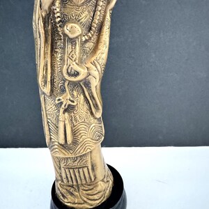 May include: A detailed bronze-colored statue of a figure, possibly a deity, with intricate carvings and a beaded necklace. The statue is mounted on a black, circular base. The figure's clothing and accessories are elaborately designed.