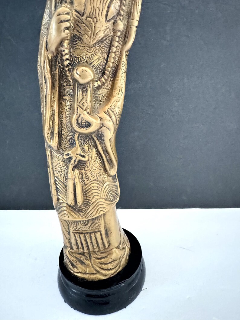 May include: A detailed bronze-colored statue of a figure, possibly a deity, with intricate carvings and a beaded necklace. The statue is mounted on a black, circular base. The figure's clothing and accessories are elaborately designed.