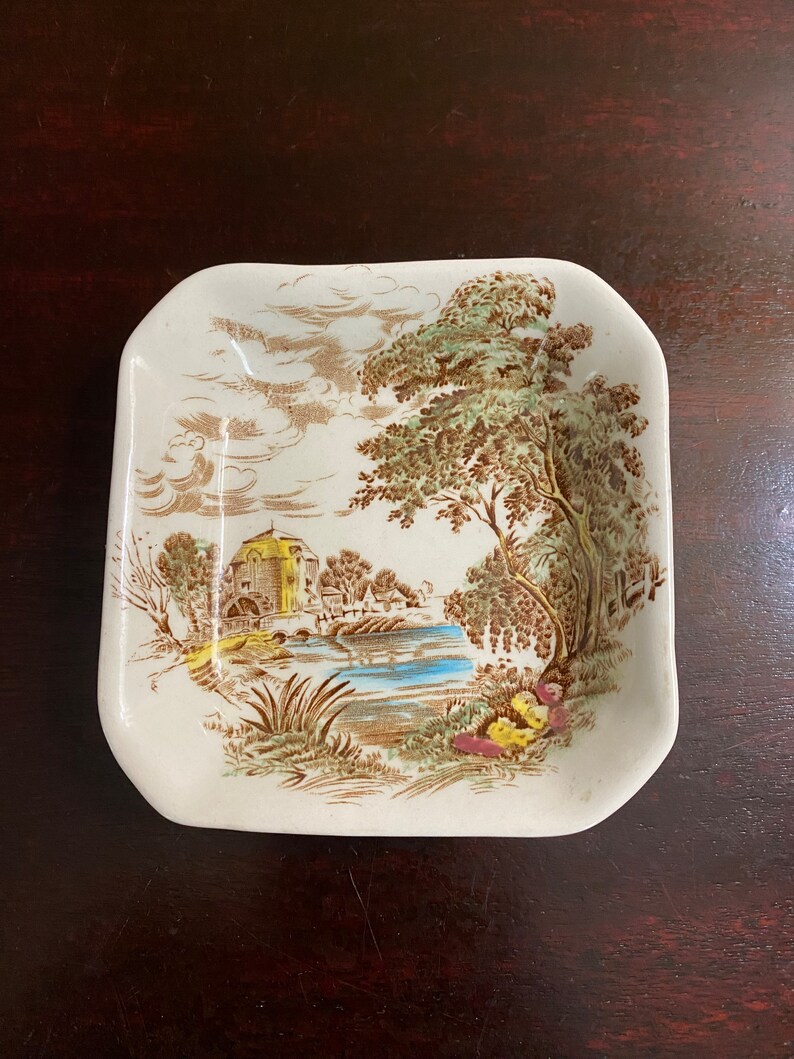 Pode incluir: Um prato quadrado de cer&acirc;mica com uma cena de paisagem marrom e verde. A cena representa um pequeno edif&iacute;cio com um telhado amarelo, um rio, &aacute;rvores e nuvens. O prato tem um fundo branco.