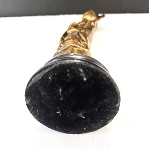 May include: A gold-colored statue of a person, mounted on a black base. The statue is detailed with visible features and is positioned at an angle, showcasing its form. The base has a soft, felt-like texture.