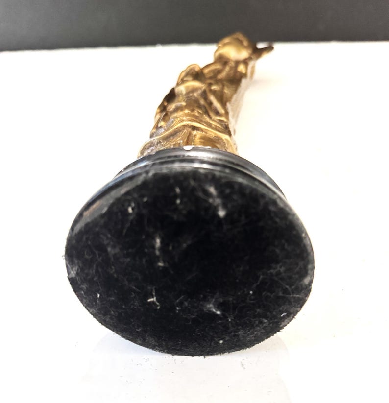 May include: A gold-colored statue of a person, mounted on a black base. The statue is detailed with visible features and is positioned at an angle, showcasing its form. The base has a soft, felt-like texture.