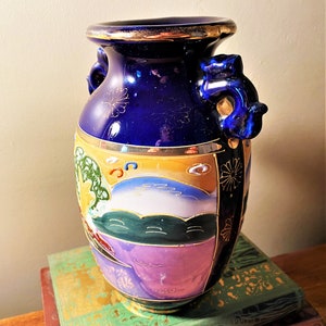 May include: A cobalt blue ceramic vase with gold trim and handles. The vase features a painted landscape scene with a mountain, trees, and a sunset. The vase is decorated with gold floral accents.