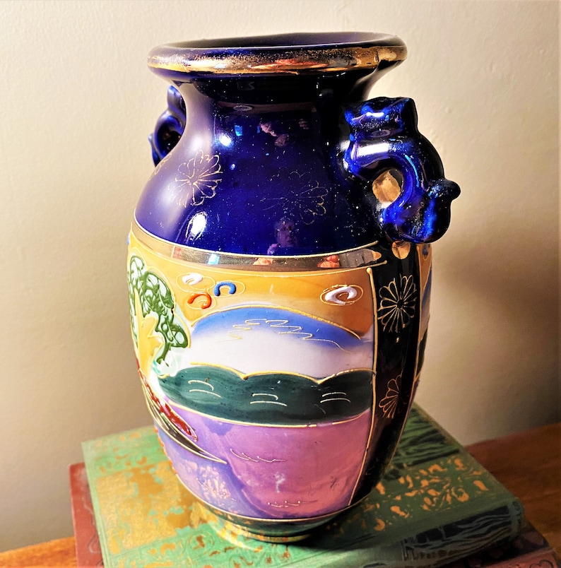 May include: A cobalt blue ceramic vase with gold trim and handles. The vase features a painted landscape scene with a mountain, trees, and a sunset. The vase is decorated with gold floral accents.