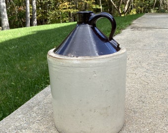 May include: A vintage brown and white stoneware crock with a handle. The crock has a brown glazed top and a white unglazed bottom.
