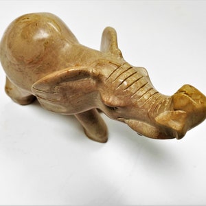 May include: A hand-carved stone elephant figurine with a light brown color. The elephant is standing with its trunk raised and its head tilted to the side.