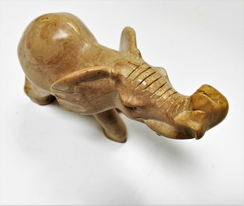 May include: A hand-carved stone elephant figurine with a light brown color. The elephant is standing with its trunk raised and its head tilted to the side.