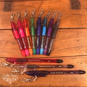 May include: A collection of colorful pens with attached keychains, arranged on a wooden surface. The pens come in various colors, including red, pink, orange, green, blue, purple, and black. The text "Mini RSVP" and "Pentel R.S.V.P." is visible on the pens.