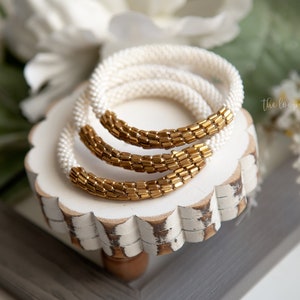 Puede incluir: Un conjunto de tres pulseras circulares de cuentas que combinan pequeñas cuentas blancas mate con cuentas cilíndricas de color oro metálico. Las pulseras están dispuestas en una formación apilada, mostrando un patrón constante.