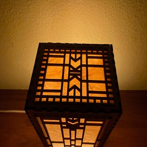 May include: A wooden lamp with a geometric design and a light brown paper shade. The lamp is lit and casting a warm glow.