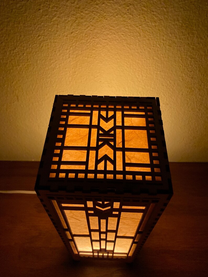 May include: A wooden lamp with a geometric design and a light brown paper shade. The lamp is lit and casting a warm glow.