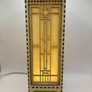 May include: A wooden table lamp with a geometric design and a light brown finish. The lamp has a rectangular base and a square shade with a geometric pattern. The shade is made of a translucent material that allows light to shine through.