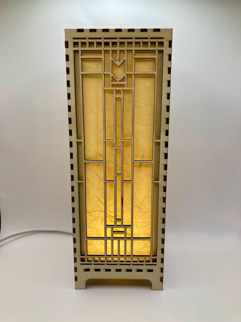 May include: A wooden table lamp with a geometric design and a light brown finish. The lamp has a rectangular base and a square shade with a geometric pattern. The shade is made of a translucent material that allows light to shine through.