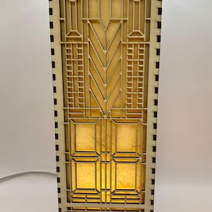 May include: A wooden lamp with a geometric design and a light brown finish. The lamp has a rectangular base and a square shade with a geometric pattern.