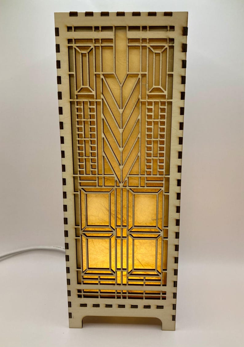 May include: A wooden lamp with a geometric design and a light brown finish. The lamp has a rectangular base and a square shade with a geometric pattern.