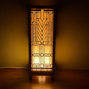 May include: A wooden table lamp with a geometric design and a warm, yellow light emanating from the inside. The lamp has a rectangular base and a tall, rectangular shade with a geometric pattern.