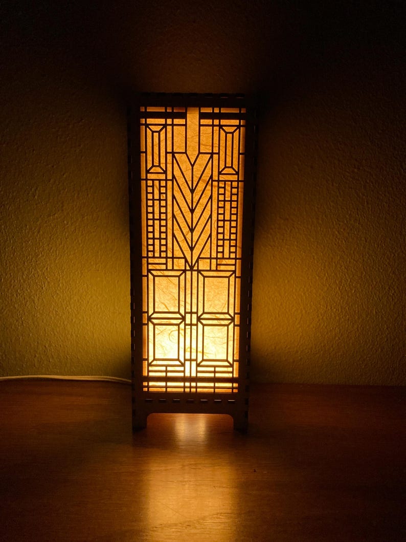 May include: A wooden table lamp with a geometric design and a warm, yellow light emanating from the inside. The lamp has a rectangular base and a tall, rectangular shade with a geometric pattern.