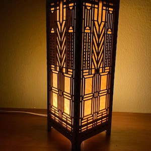 May include: A wooden lamp with a geometric design and a warm, yellow light emanating from the inside. The lamp is on a wooden surface.